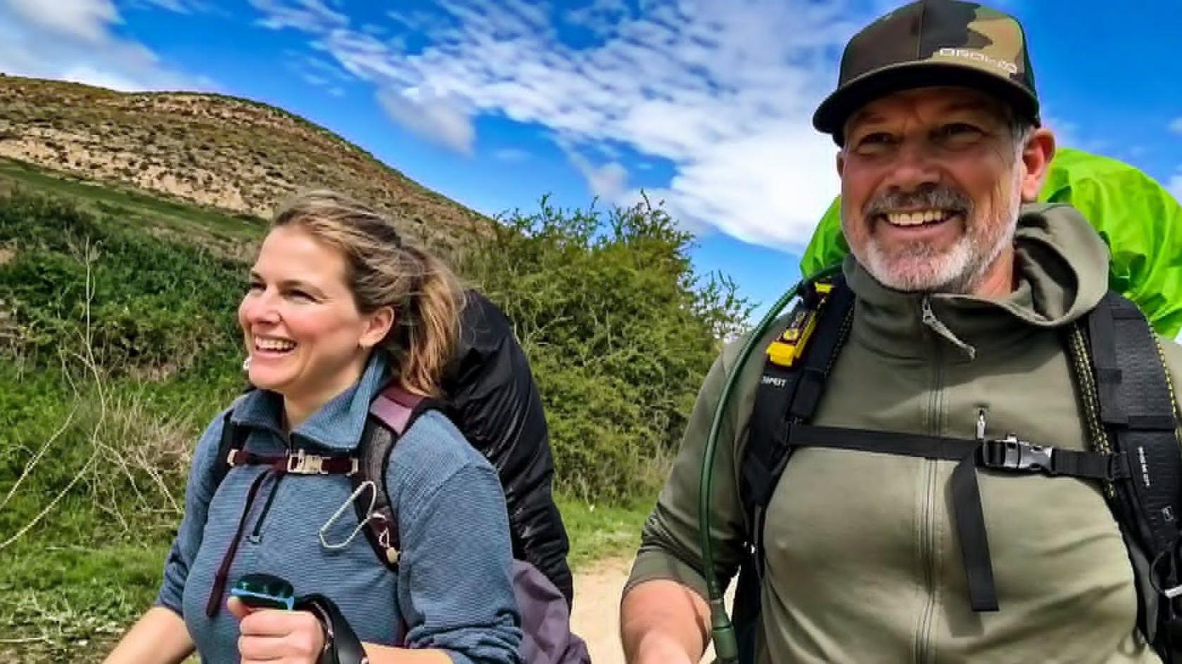 An image showing Christie side by side with her partner Steve mid-hike. They are both smiling and wearing outdoorsy gear, including hiking backpacks. Behind them is a blue sky and grass-covered hills.