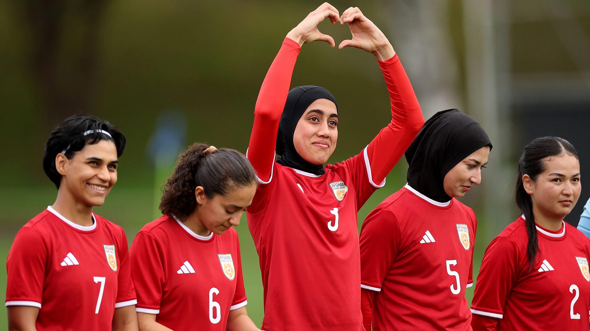 Five women in red football kit stand in a line. The one in the middle is holding her arms above her head and her hands form the shape of a heart.