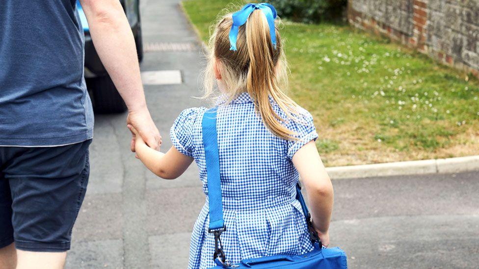 A blonde girl in chequered blue school uniform dress holds her father's hand as she walks down a pavement