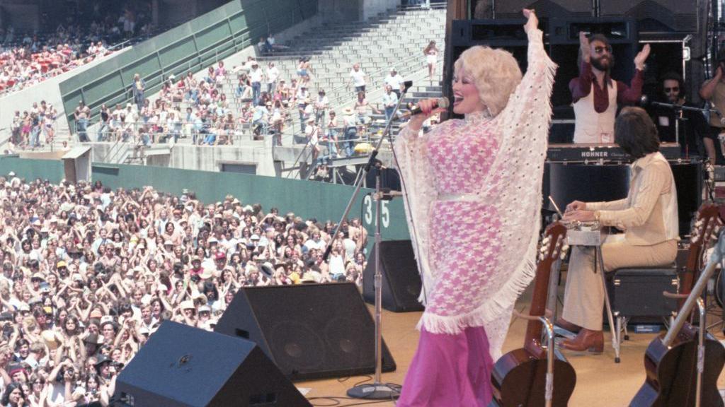 A side view of Parton singing on stage, and you can see the huge crowd watching her.
