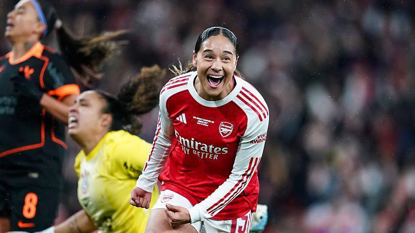 Olivia Smith has a happy expression on her face as she celebrates scoring for Arsenal in the Women's Champions Cup 2026 final against Corinthians. Two anguished Corinthians players are behind her, one in largely black kit and the other in a yellow goalkeeper shirt, while Smith wears a long-sleeved variant of Arsenal's traditional red and white shirt.
