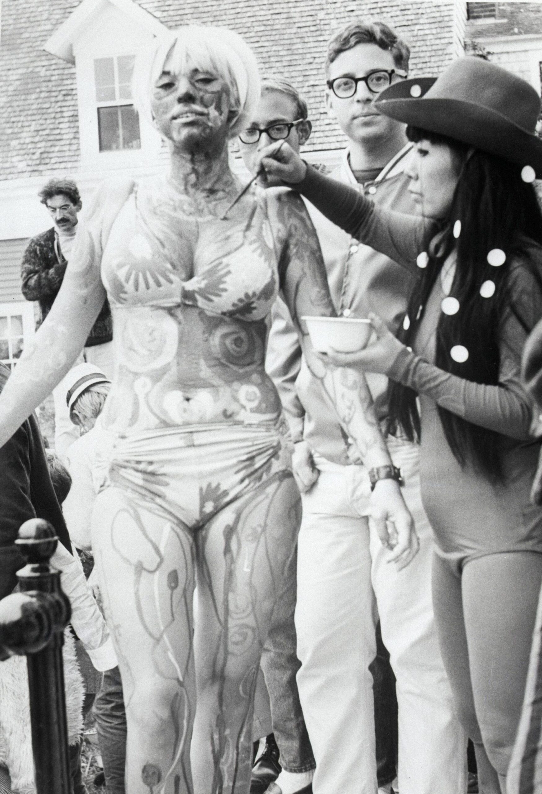 Black & white photo of Yayoi Kusama in 1967 painting a young woman wearing a bikini at a Body Festival in Provincetown, Massachusetts.