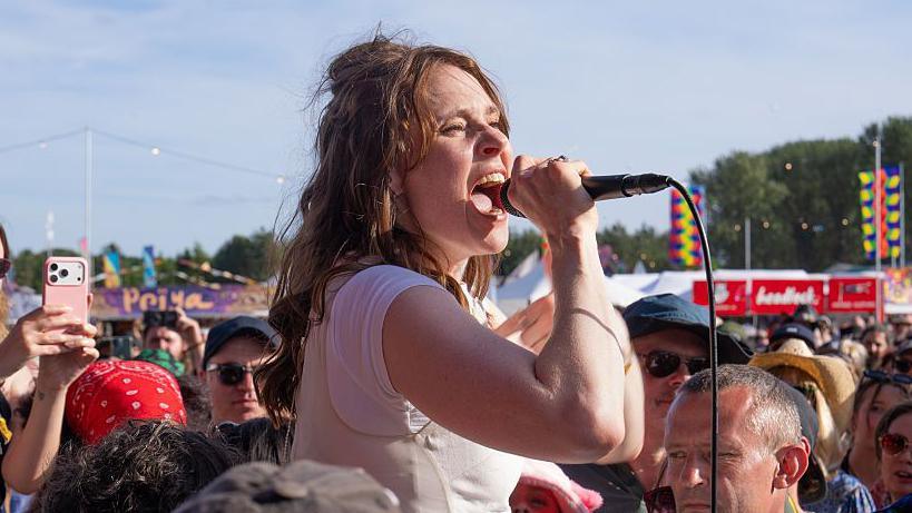 Kate Nash pictured in a white top singing into a handheld microphone while standing among a large crowd at an outdoor music festival. Audience members surround the performer, with some filming on smartphones. Colourful festival banners, food stalls and a line of trees are visible in the background under a bright blue sky.