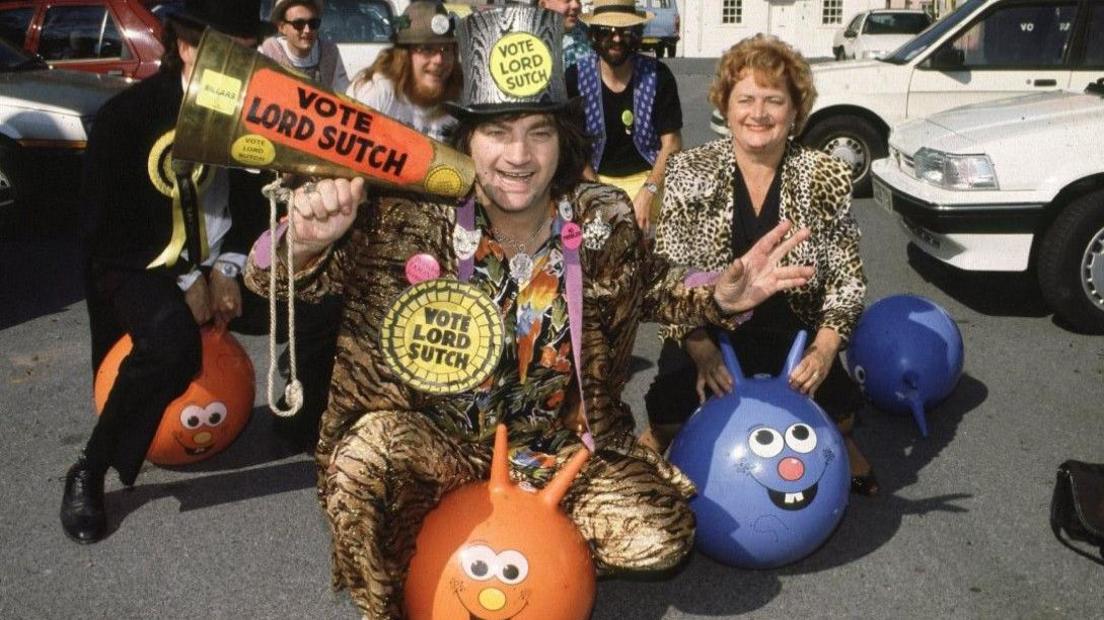 Screaming Lord Sutch of the Monster Raving Loony Party, at their 1990 party conference. He wears a leapoard skin suit with a matching leopardskin top hat and is sitting astride an orange spacehopper. He is wearing several giant yellow and black rosettes and carries a golden loudhailer, all with the slogan Vote Lord Sutch. He is surrounded by a group of supporters also riding spacehoppers in a car park.