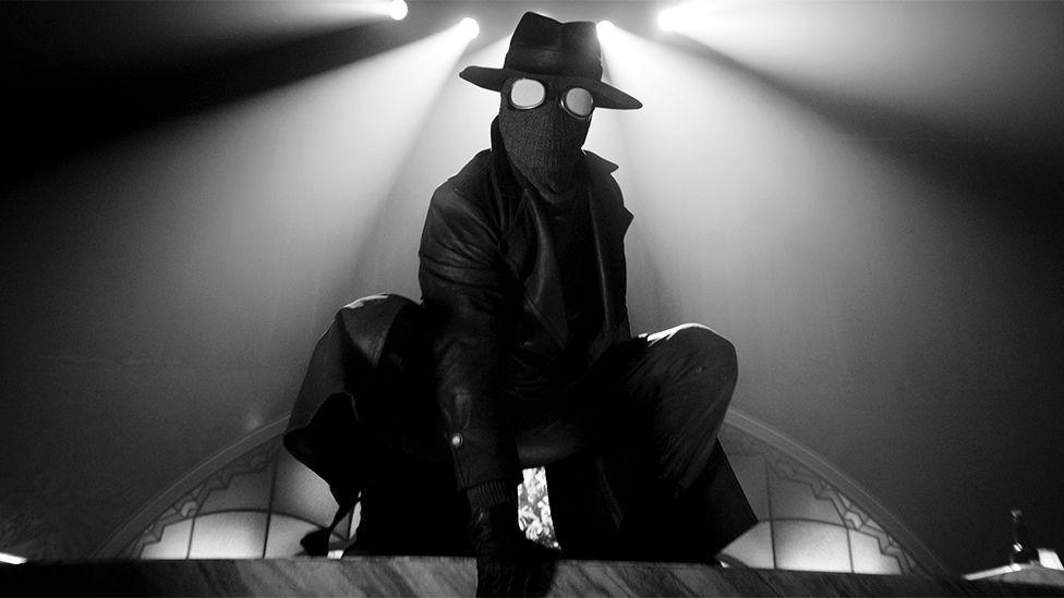 A black and white image of a character called The Spider, wearing a round hat and mask, with a leather jacket, with a white spotlight above him.