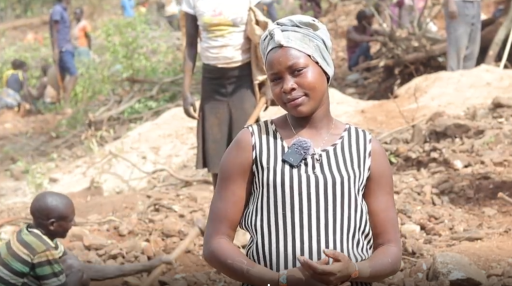 Abigael Chelengat in a grey head wrap and black and white striped vest top looks at the camera with her head tilted to one side. Behind her people can be seen standing around among the earth and rocks.