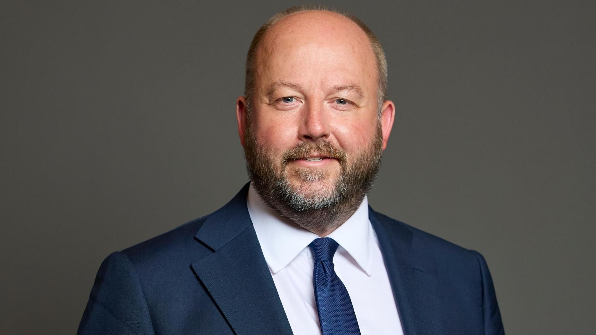 A portrait image on a grey background of Nick Timothy wearing a blue suit, white shirt and tie.