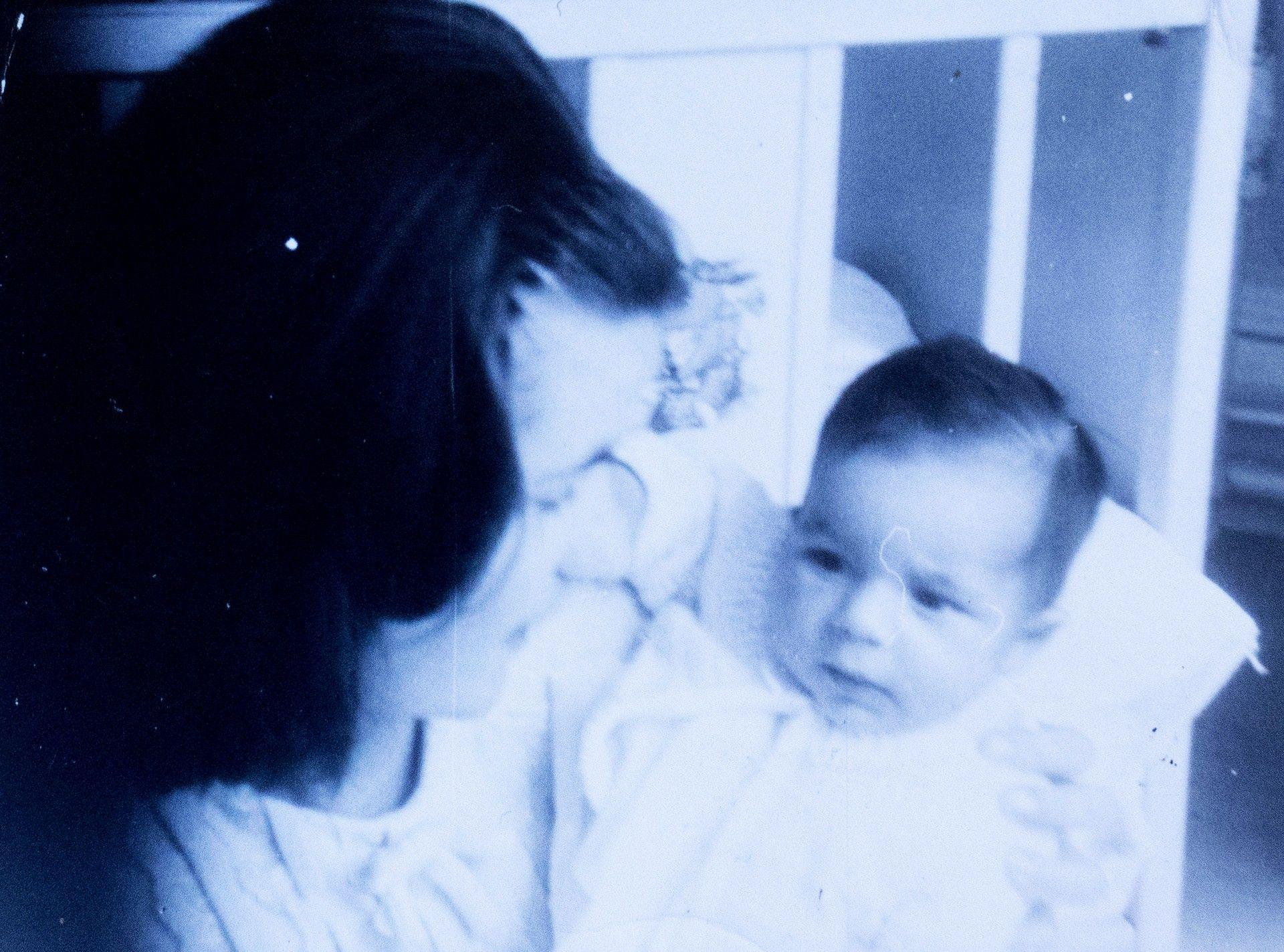 An old,  black and white photograph of a young woman cradling a baby. They are crouched next to a cot.