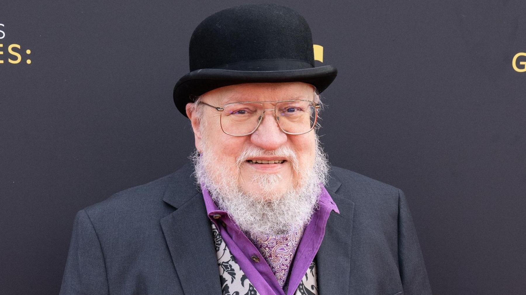 George RR Martin with grey beard, glasses, bowler hat, purple shirt, patterned waistcoat and grey suit jacket, looking at the camera and smiling.