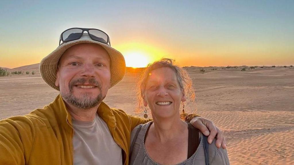 A selfie taken by Nick Smith showing him wearing a yellow jacket and a beige undershirt, and a beige hat with sunglasses sitting on top of his head, his arm around his wife, who is smiling and has an updo and is wearing dangling earrings. Behind them is the sun setting over the Moroccan desert.