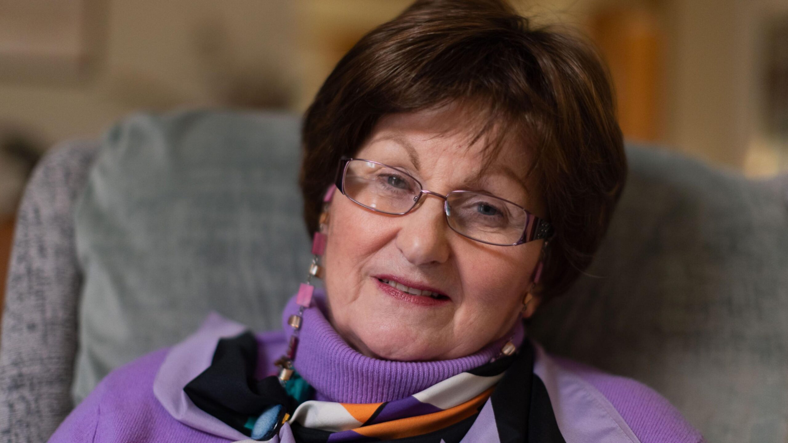 An elderly woman with short dark hair. She is wearing glasses, a purple jumper and a colourful scarf. She is sitting in a grey armchair, smiling at the camera.