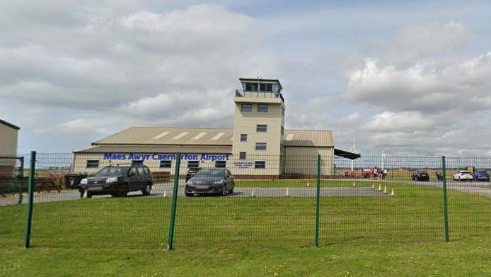 A small cream building airport. It has a watchtower and surrounded by green grass and fields. There's a car park with multiple cars around the building.