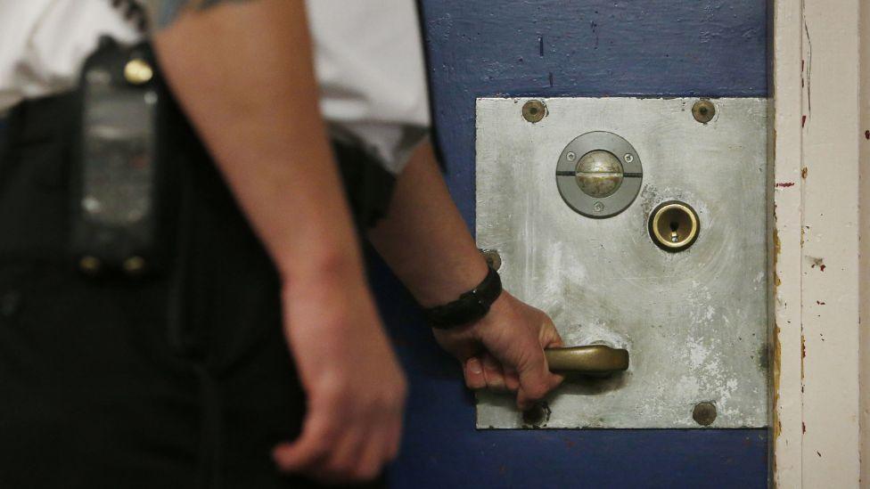 A prison officer with his left hand on a cell door. His upper and lower body is out of shot.