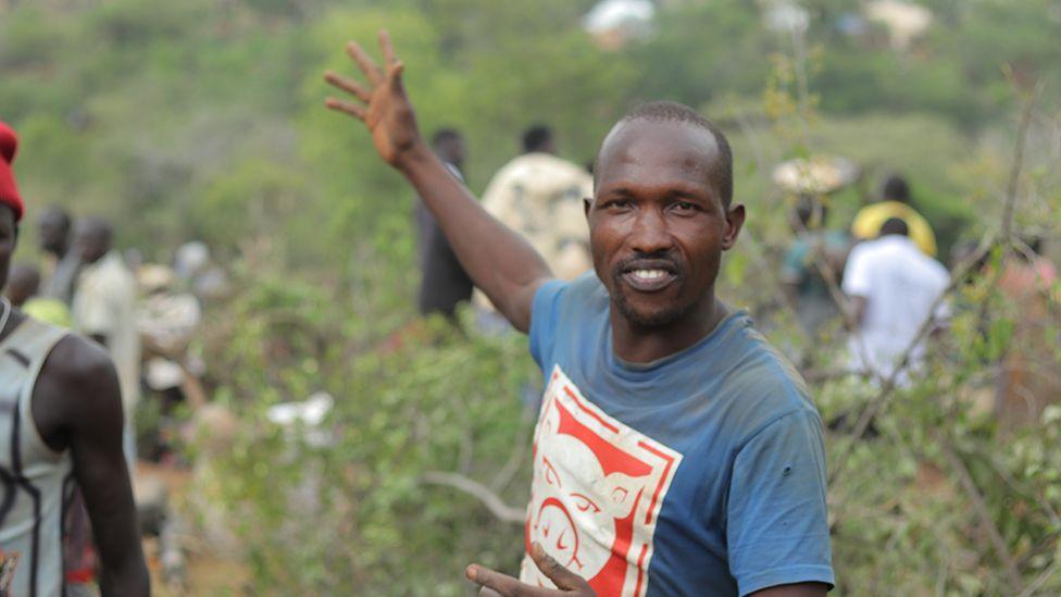 Manasse Lomachar in a blue T-shirt with a cartoon pig printed on it looks at the camera but gestures behind him with his right hand. In the background, out of focus, people can be seen working among the shrubs.