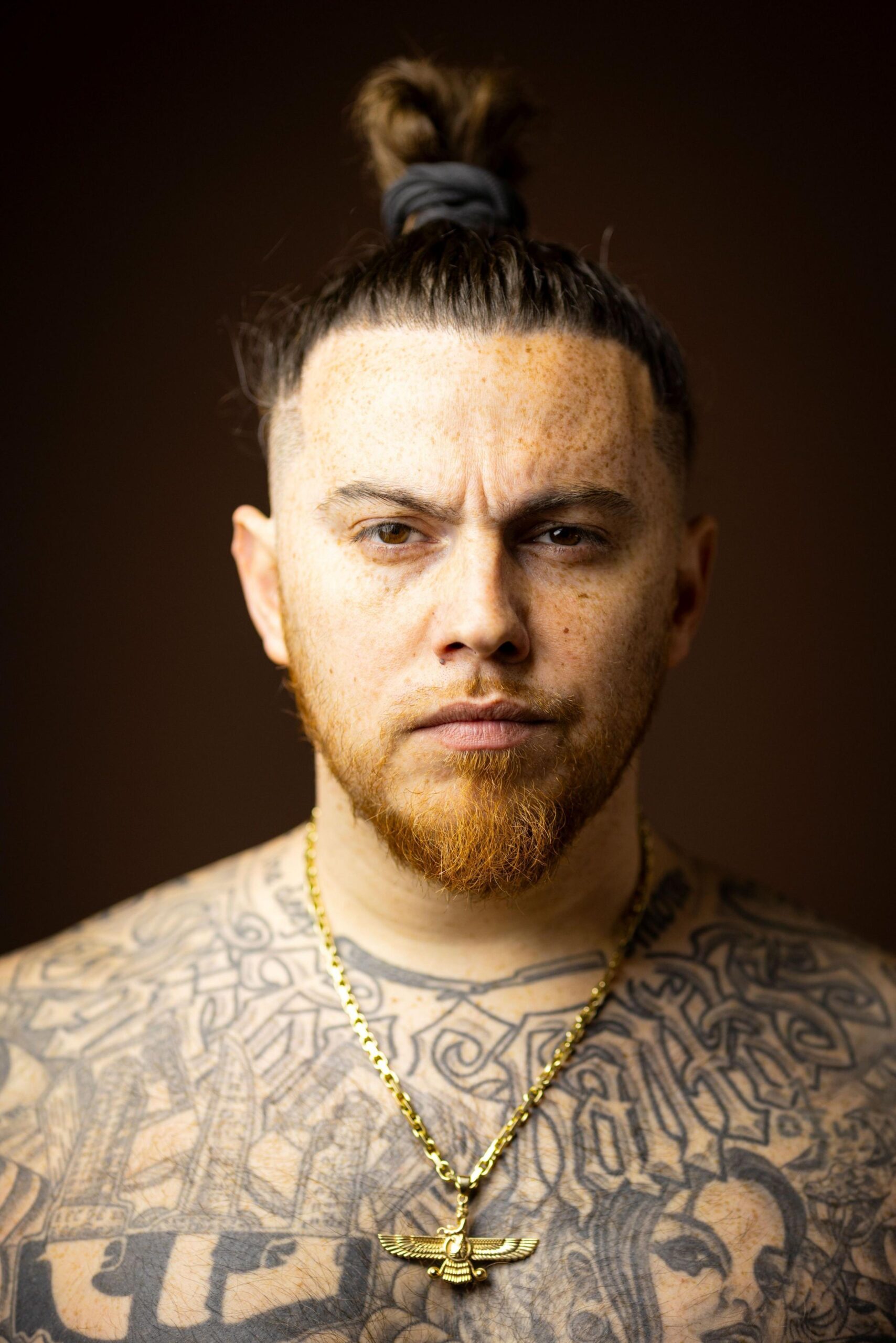 Close-up studio portrait of a person with hair tied in a top knot, freckles, a short beard, and extensive black-and-grey tattoos across the chest and shoulders. The person wears a gold chain necklace with a winged pendant and faces the camera against a dark brown blurred background.