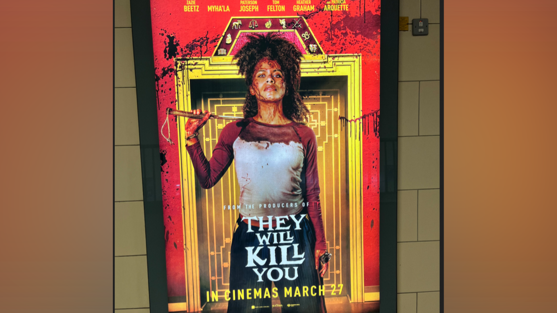 A film poster on an underground station wall shows a blood-splattered woman standing in front of an ornate doorway. She wears a blood-stained shirt, has blood splattered across her face, and holds a weapon resting over her shoulder. Red blood splatters cover the background behind her. The text reads: "From the producers of They Will Kill You. In cinemas March 27."