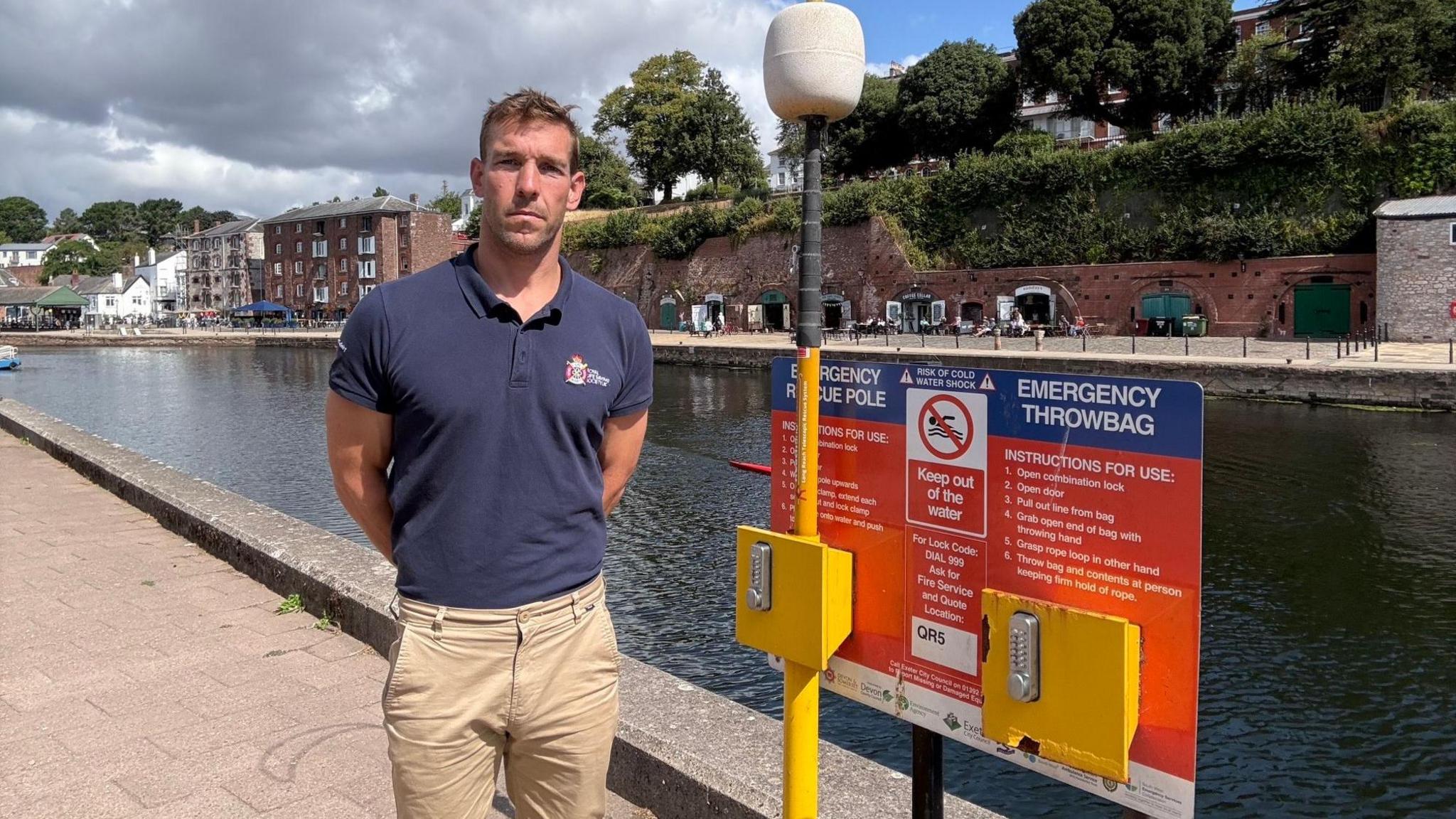 A man wearing a blue polo shirt and brown trousers is stood next to a yellow, red and blue safety sign. Both the man and the sign are stood infront of a river. There are some houses and trees in the background, and the sky is blue with some clouds.