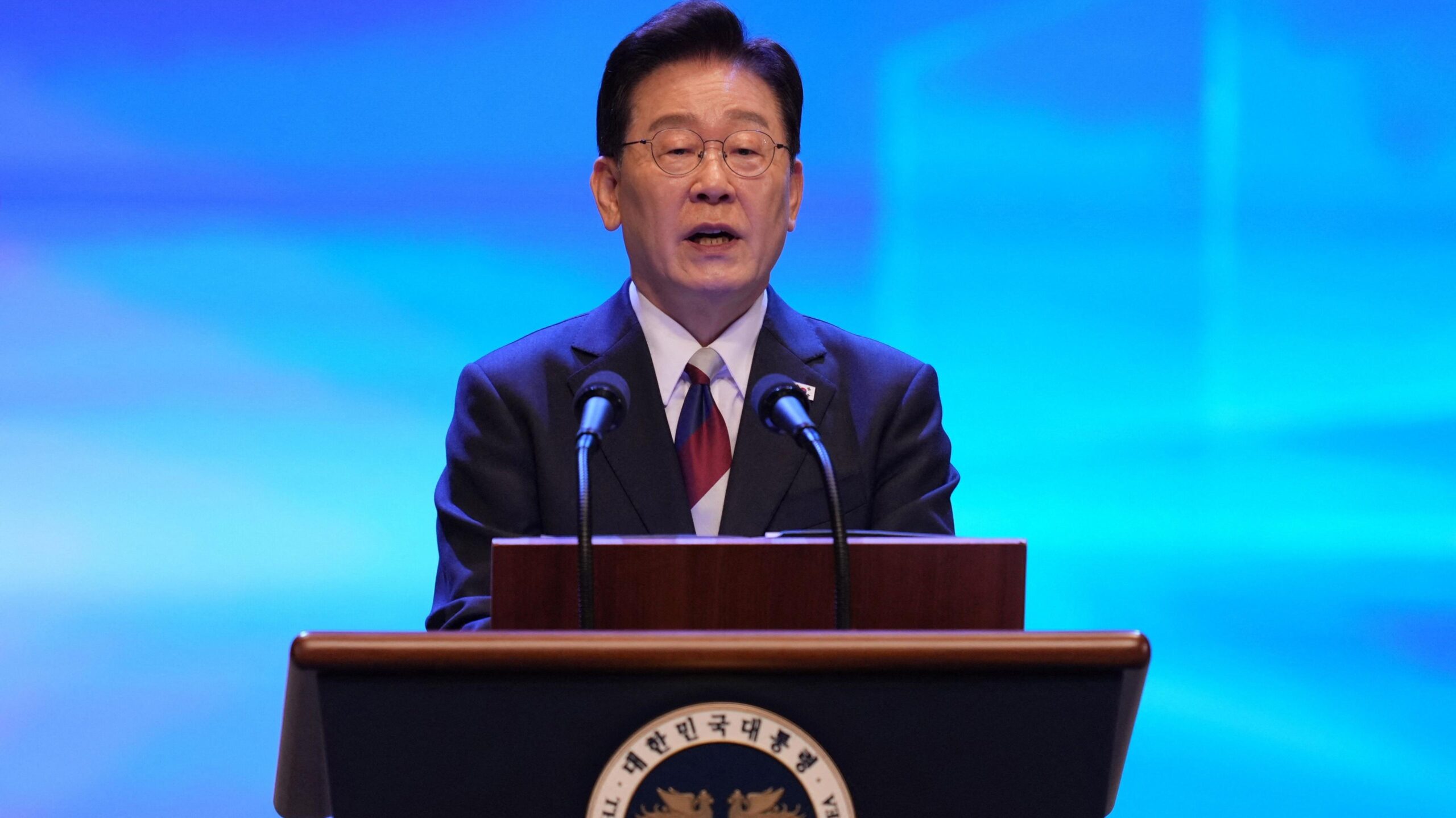 Lee Jae Myung speaks at a lectern during a Liberation Day ceremony. He wears a dark suit and glasses, in front of a blue backdrop.