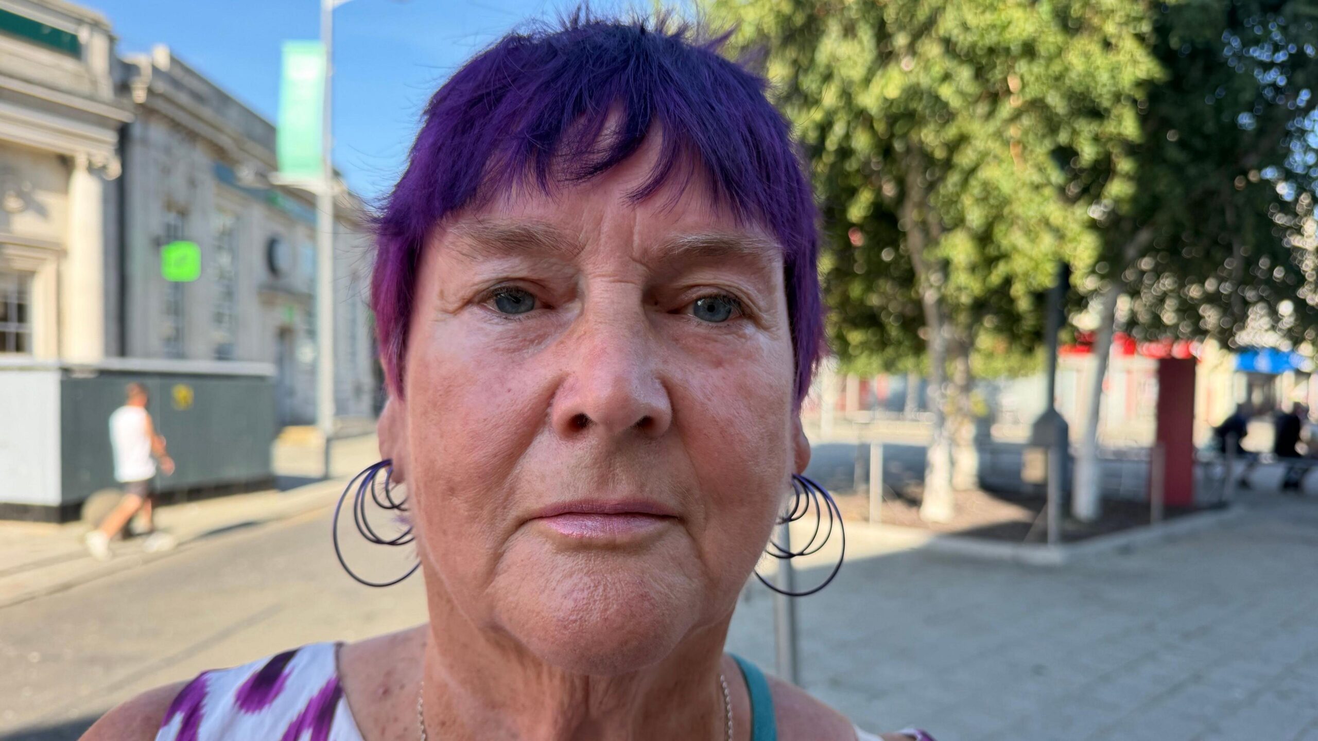 Mary Williams has short purple hair. She is wearing a white and purple vest top in Clacton town centre and large hoop earrings.