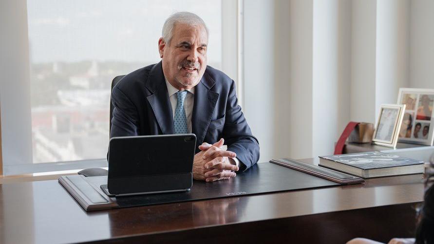 A man with grey hair, wearing a navy suit, sat at a wooden desk against a window with an ipad