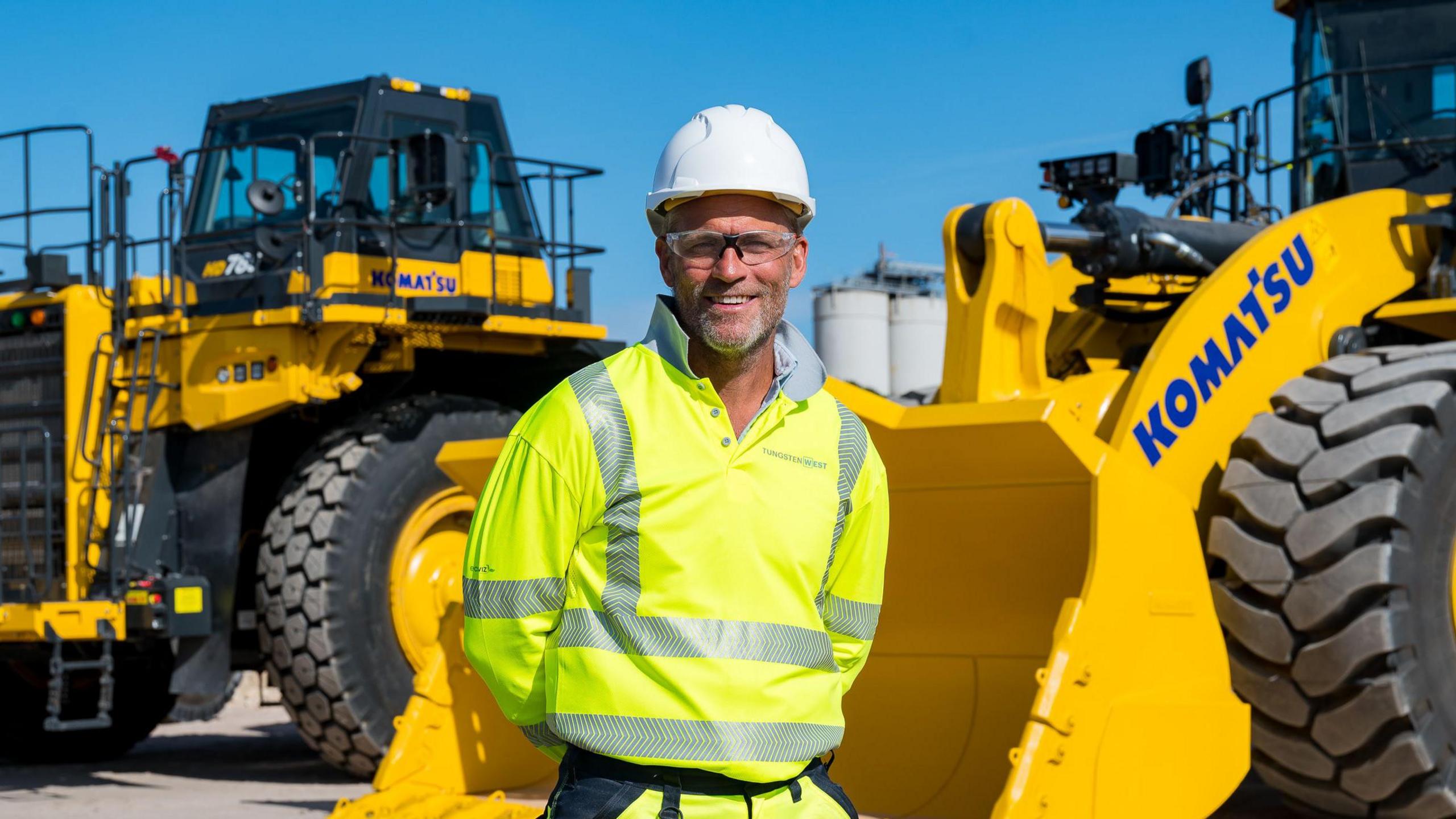 Jeff Court smiling at the camera with a yellow hi-vis. He's stood in front of a large digger.
