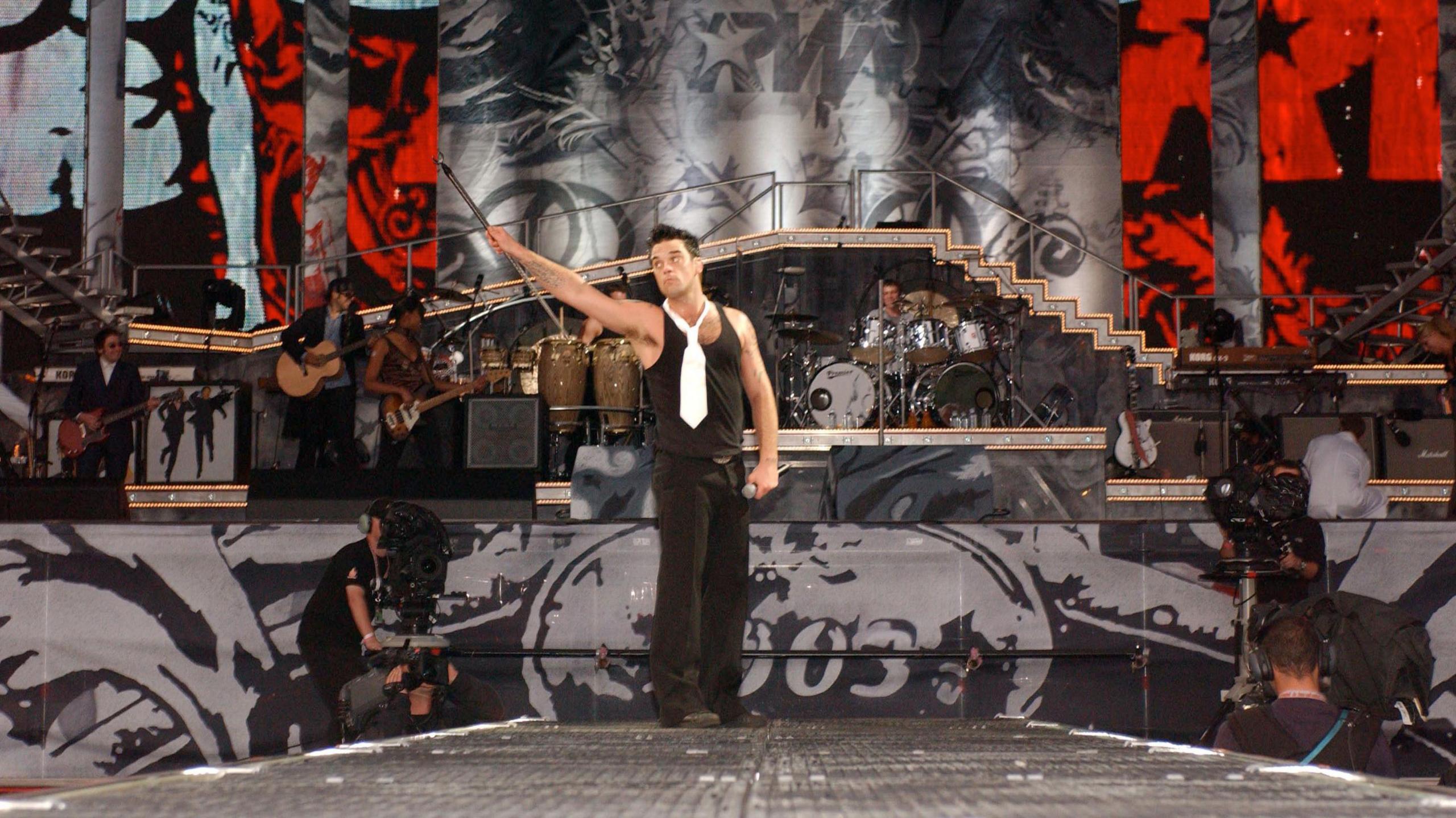 Robbie Williams standing on stage at Knebworth holding a mic stand in one hand