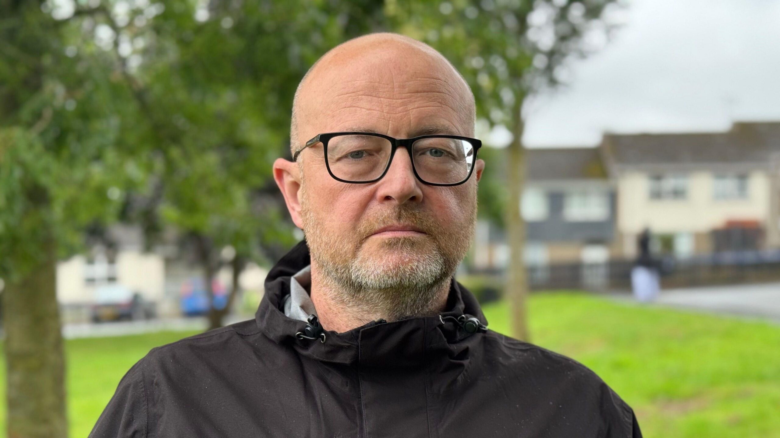 A male with a bald head, glasses and a beard. He is wearing a black rain jacket.