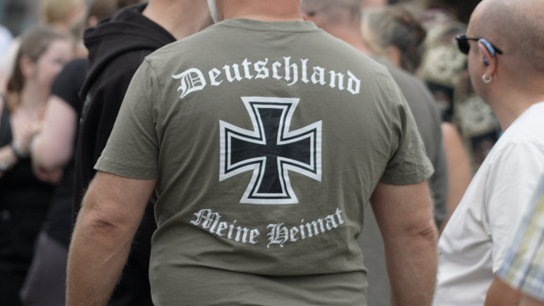 The black of a man wearing a khaki green t-shirt which says 'Deutschland, Meine Heimat' written in a Gothic-style font. The t-shirt shows the insignia of the iron cross
