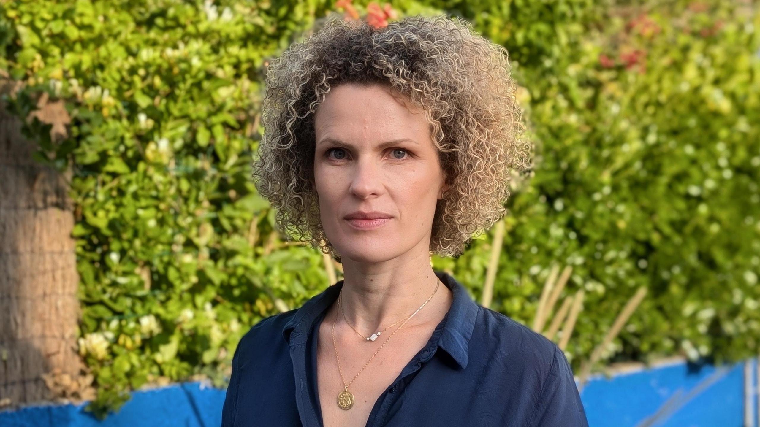AnaĂ¯s de Vos is wearing a dark blue shirt with an open neck and is standing in front of bushes and a low, bright blue wall. She is wearing a gold necklace and has light, curly hair.