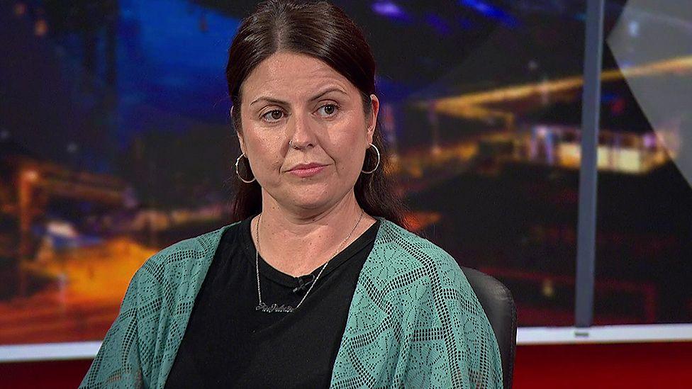 Michaela Rafferty is wearing a dark top with a green cardigan over it. She has dark hair, is wearing large hoop earrings and a silver necklace. She is pictured in a television studio.