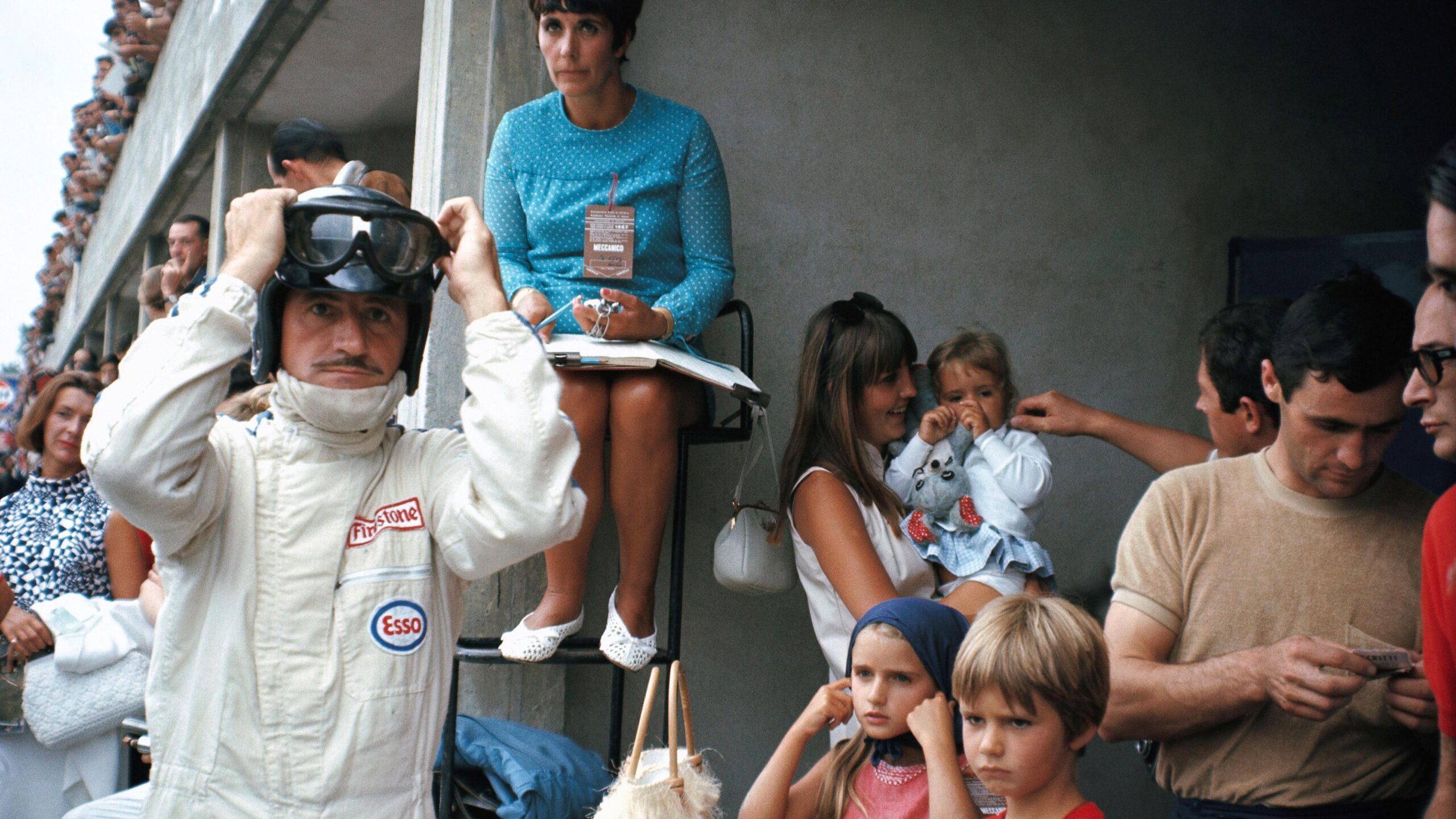 Graham Hill adjusts his race helmet and visor in the pits before the 1967 Italian Grand Prix. Gathered around him is his family, including wife Bette and his children, including Damon Hill, who is stood at the front and was then aged six