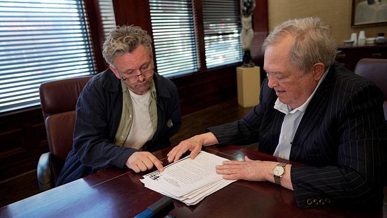 A meeting takes place around a polished wooden boardroom table, where Sheen and Stewart sit side by side reviewing a printed report. Stewart, who has grey hair and is wearing a pinstripe suit points to text on the top page while Sheen rests a hand on the document. Sheen is wearing a navy overshirt over a green shirt and white T-shirt. Large windows with partially closed blinds line the wall behind them, and office furnishings and artwork can be seen in the background.