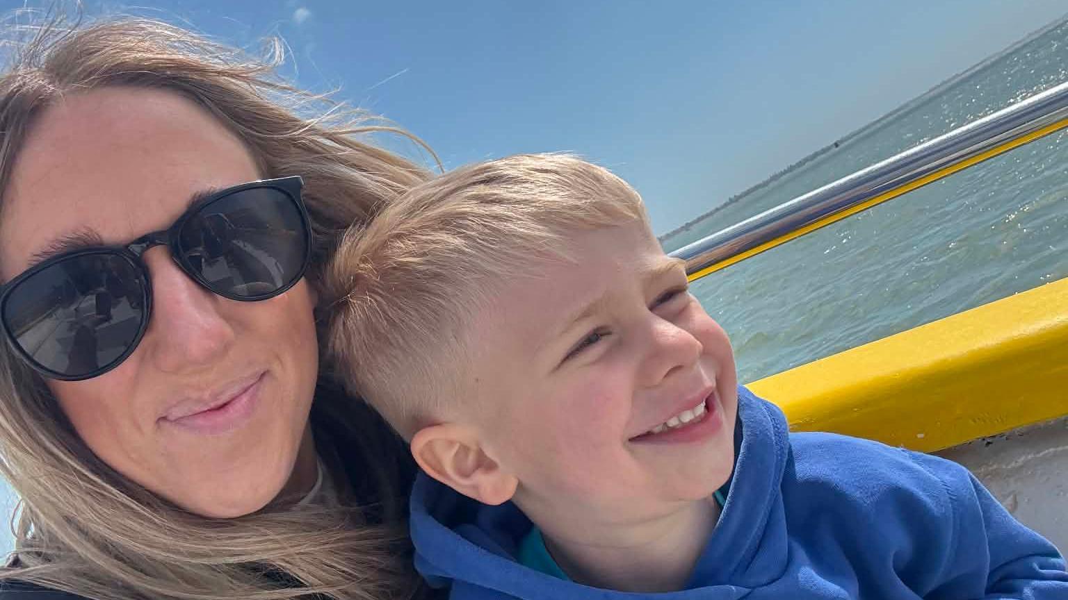 Jade and Reggie smile as they sit on a boat with the sea in the background. Jade is wearing black sunglasses and has long blonde hair. Reggie is wearing a blue jumper and has blonde hair.