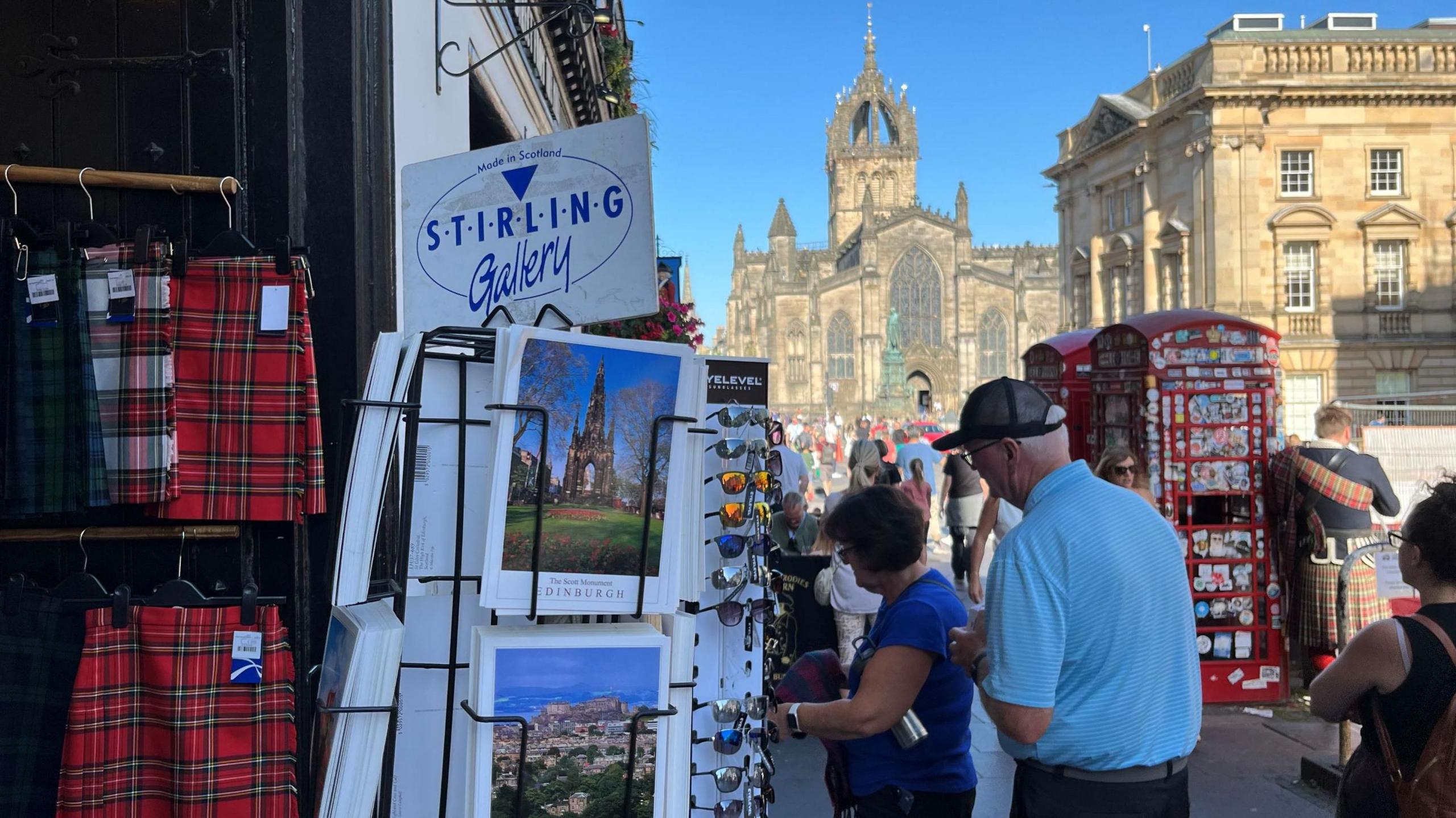 In the foreground there are postcards of Edinburgh and red tartan kilts with a man and a woman looking through other items in the gift shop. In the background is a traditional red telephone box, a man in full highland dress and the spires of St Giles Cathedral.
