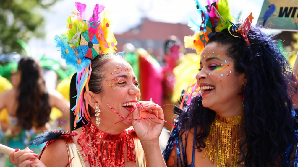 Two women with glitter on their faces and colourful headpieces smile at each other.
