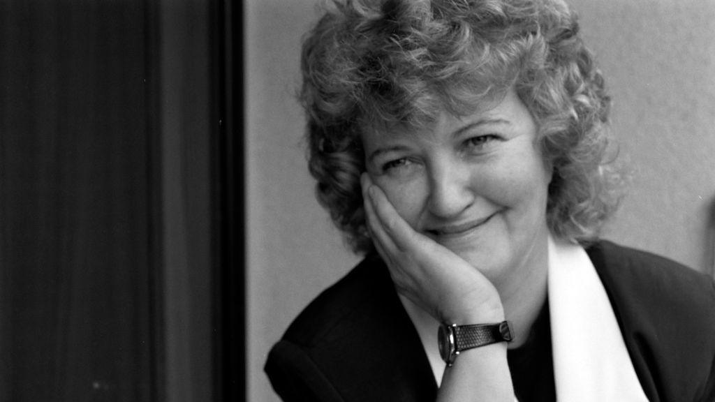 A black and white photo shows a woman with short curly hair. Her right hands holds her right cheek, which has a watch on its wrist. She has on a dark top and white scarf around her neck.