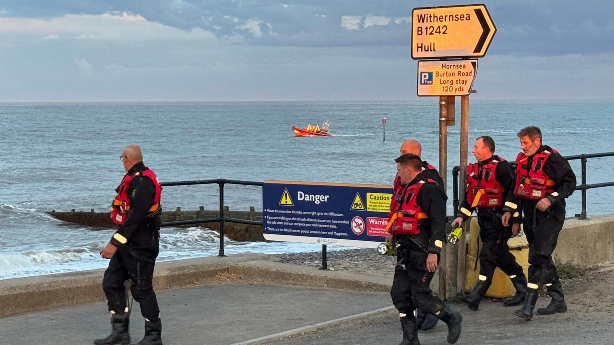 Five men wearing black suits and red lifejackets are walking along the seafront. Some are looking out to sea, some are looking ahead. The sea is visible behind them. An orange search and rescue boat is visible in the distance.