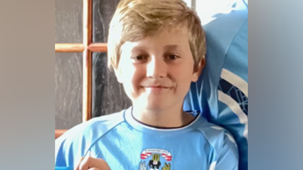 A young boy with short blonde hair, parted on the left side of his head, looks into the camera. He wears a blue football top with the top of a club logo visible just at the edge of the shot. He stands in front of a glass-paned door with a brown wooden frame.