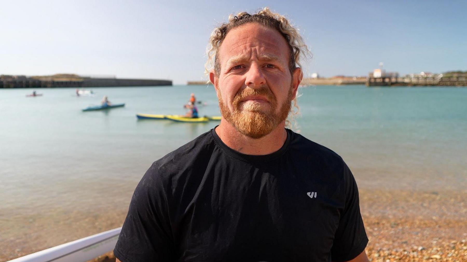 Wesley Horscroft pictured on Kingston Beach, Shoreham. He has blondish hair and a ginger beard. He is wearing a black t-shirt. Paddleboarders can be seen in the background.