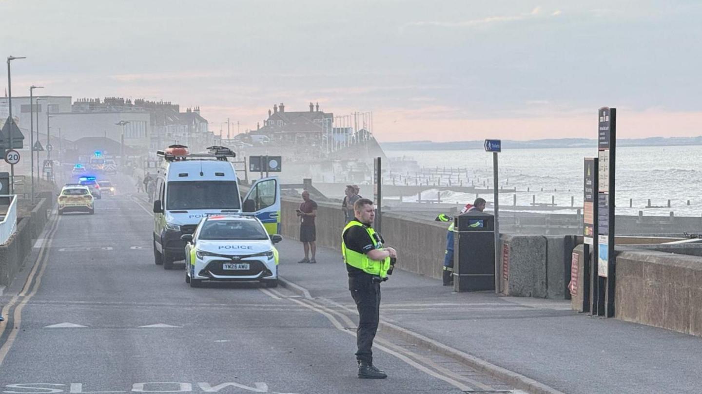 Emergency vehicles along the seafront and an officer standing in the foreground. There are a number of buildings in the background and blue flashing lights can be seen.