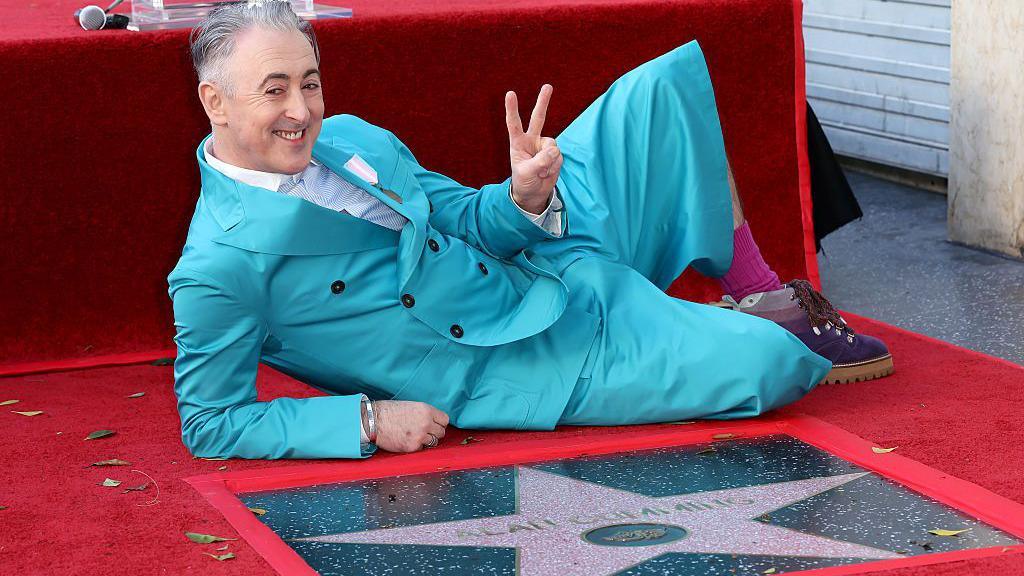 Alan Cumming, wearing a turquoise suit, poses on the ground behind his Hollywood Walk of Fame star. He is on one side, supported by one elbow, with the other hand raised in a two-finger peace sign.