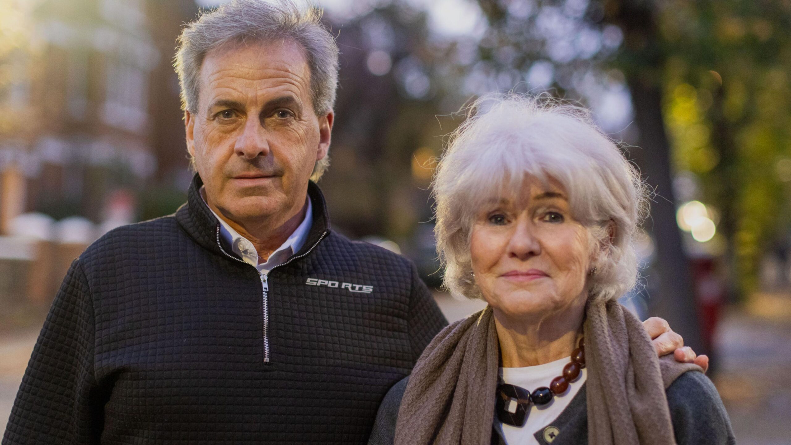 A man and a woman looking at the camera. The man is on the right and has short grey hair and has his hand on the woman's left shoulder. The woman has chin-length grey hair and is wearing a scarf and a large necklace.