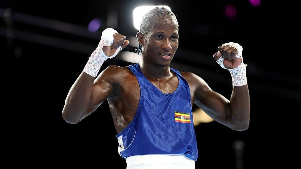 Nuhu Batte, a boxer wearing a blue top with a small Ugandan flag printed on it, holds his fists up in the air while standing in a boxing ring.
