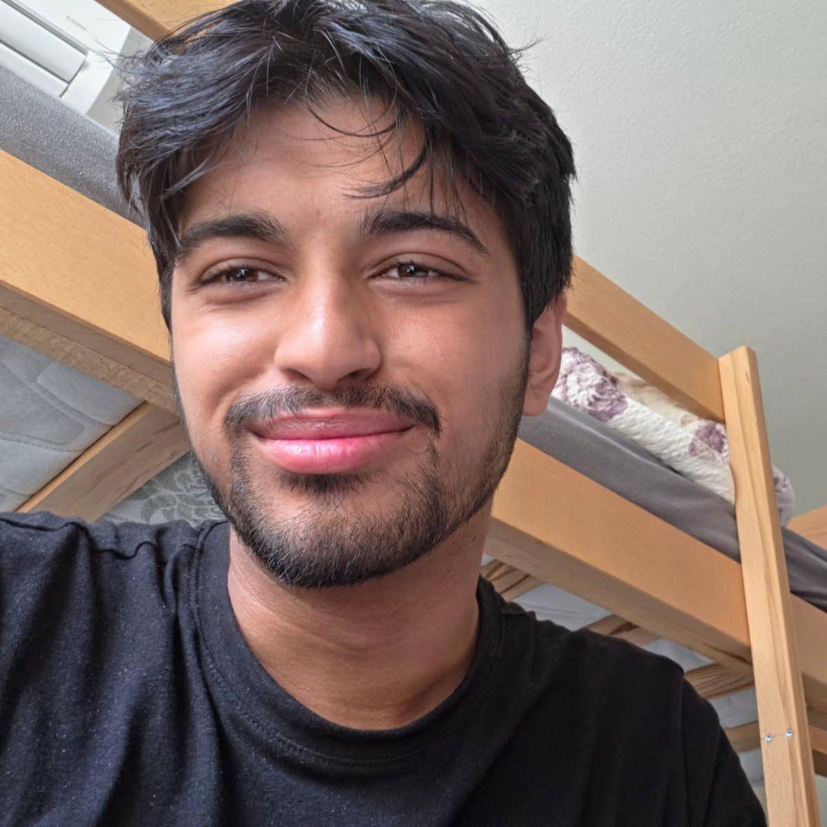 A man with black hair and a black beard smiling, he is wearing a black top and behind him is wooden bunk beds.