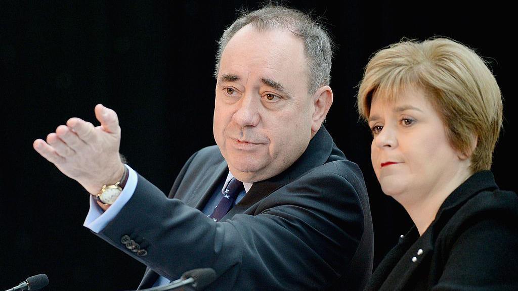 Alex Salmond, left, wearing a dark suit and looking away from the camera, raising his left armd and hand in the air. Nicola Sturgeon, right, wearing a black top. Both are sitting against a black background.
