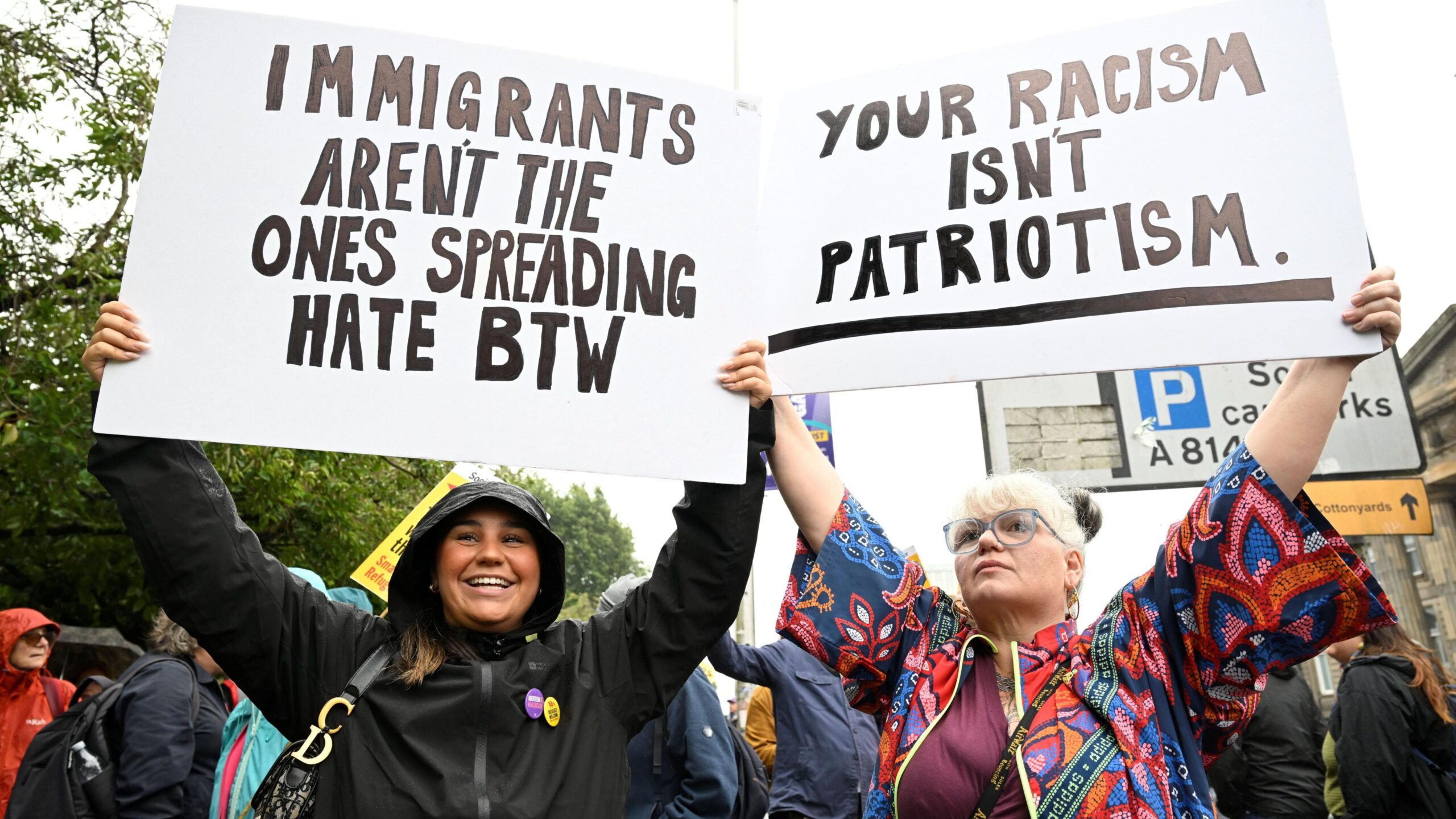 People at a public demonstration hold large placards above their heads. One sign reads, “IMMIGRANTS AREN’T THE ONES SPREADING HATE BTW,” and another reads, “YOUR RACISM ISN’T PATRIOTISM.” Other demonstrators, trees, and road signs are visible in the background.