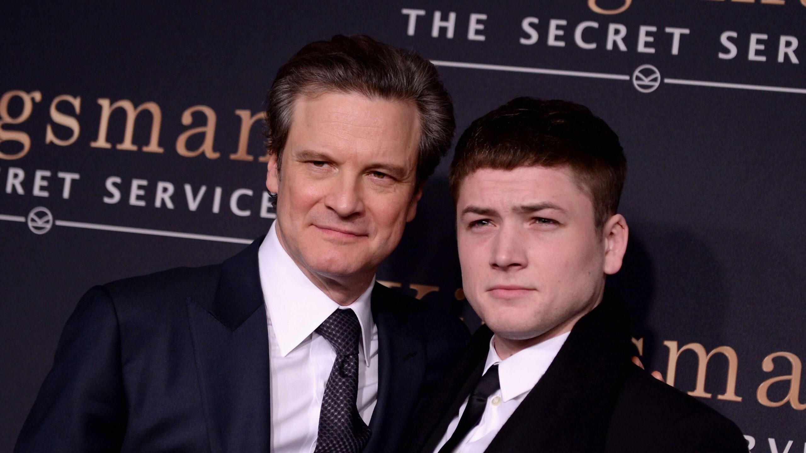 The actors stand next to each other on the red carpet, with a black background with the film title on it.  They wear dark suits with white shirts and dark ties.