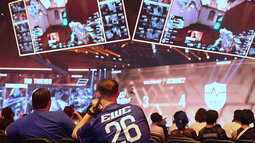 Spectators are shown in an arena looking at teams competing during the 2026 Esports World Cup in Paris. Four large screens can be seen showing the action taking place.