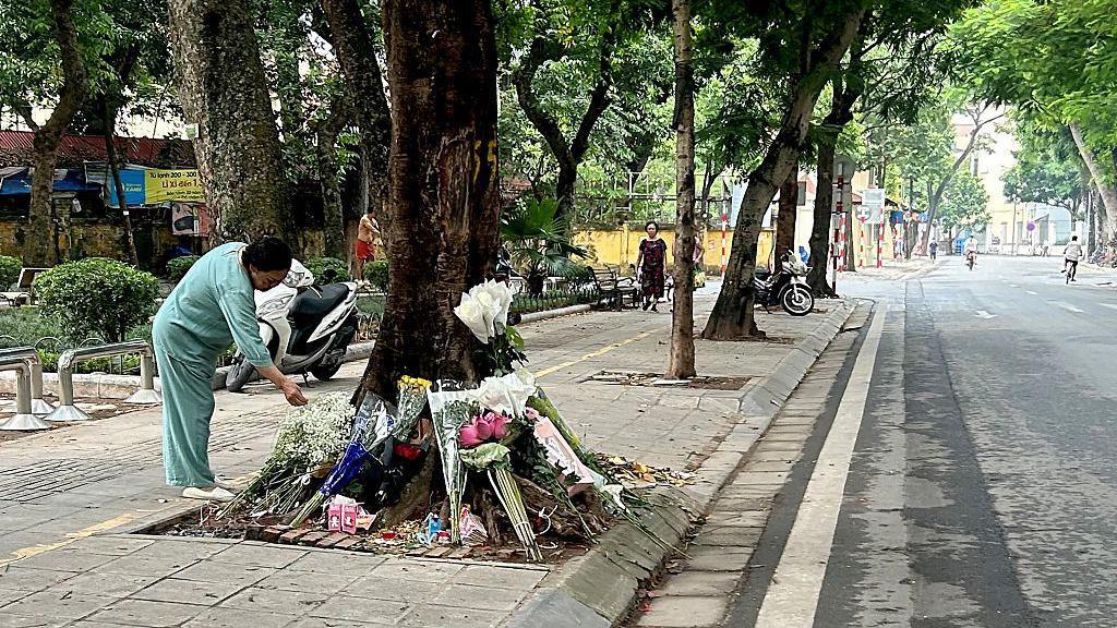 A woman in mint green clothes bends over a ring of flower bouquets arranged around the trunk of a roadside tree