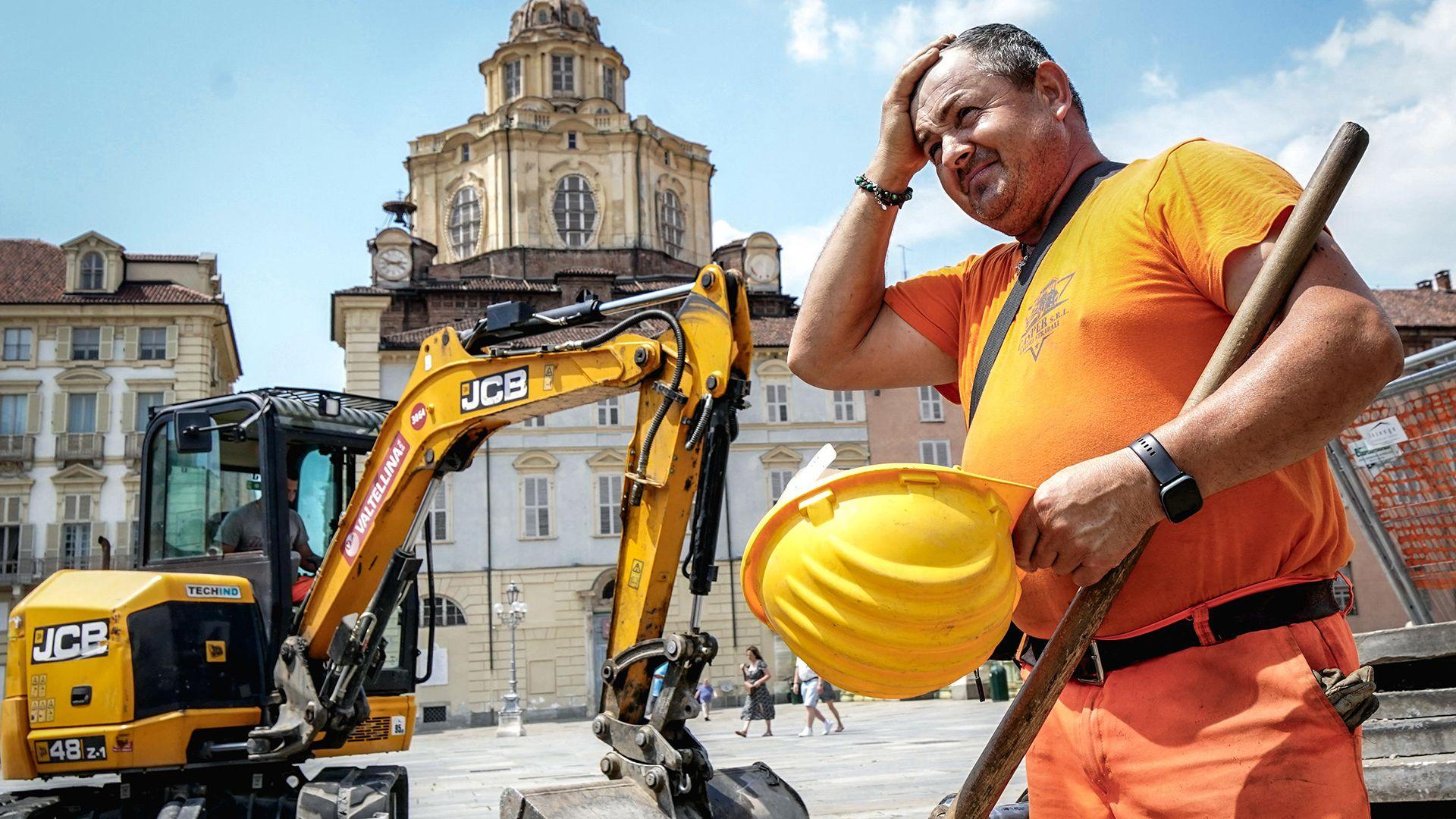 A construction worker wearing orange hi vis uniform holds a yellow construction helmet and wipes his head in front of a JCB digger next to a public square in Turin, Italy on 25 June.